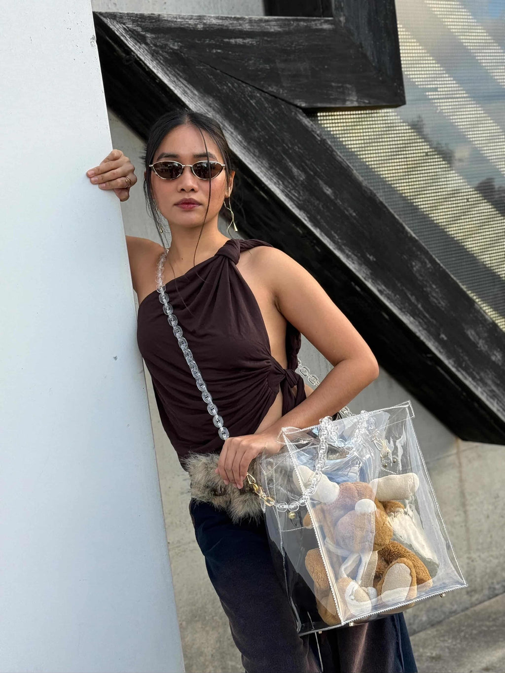 Woman holding a clear handbag with stuffed animals, standing against a building.