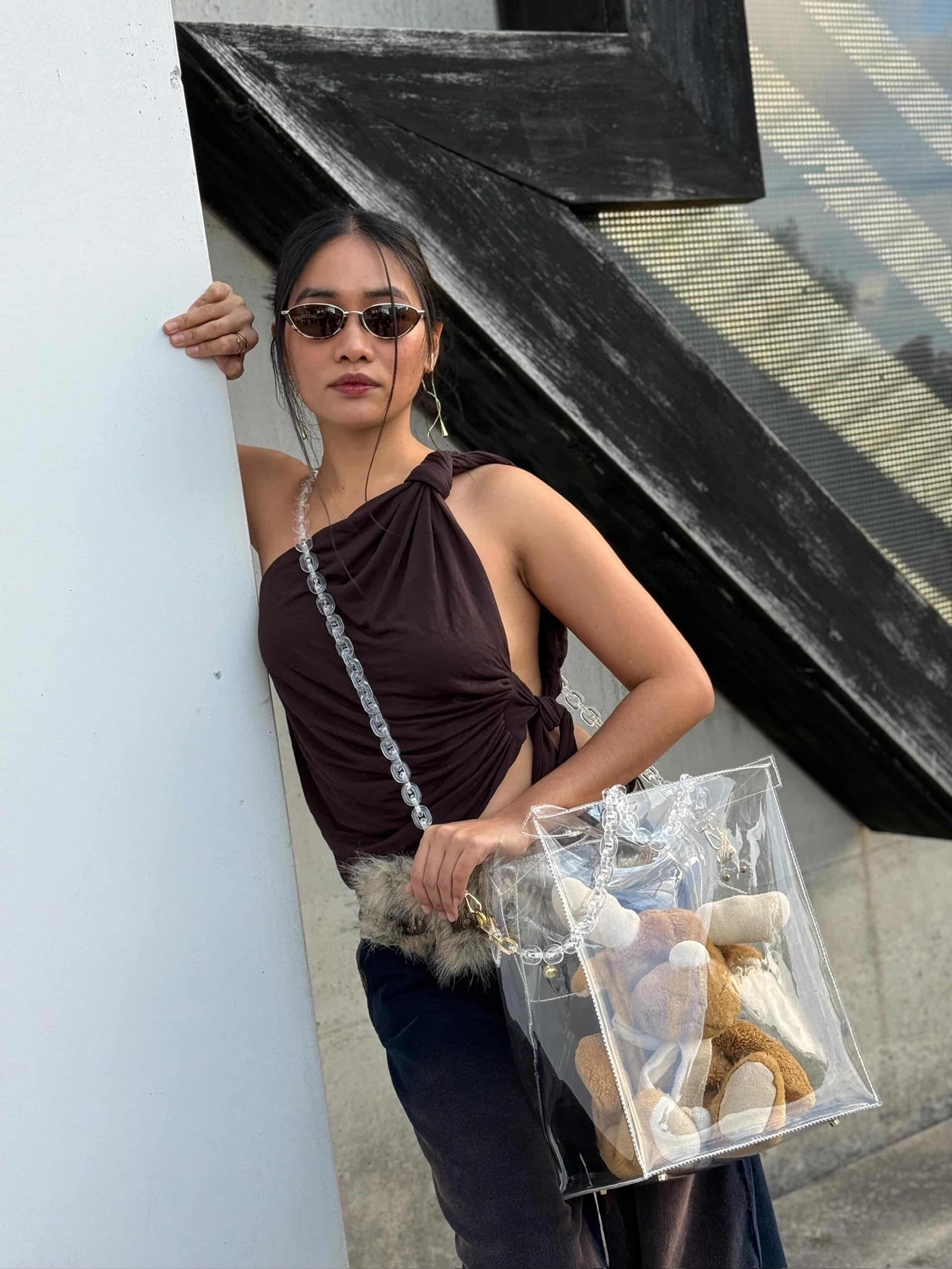 Woman holding a clear handbag with stuffed animals, standing against a building.
