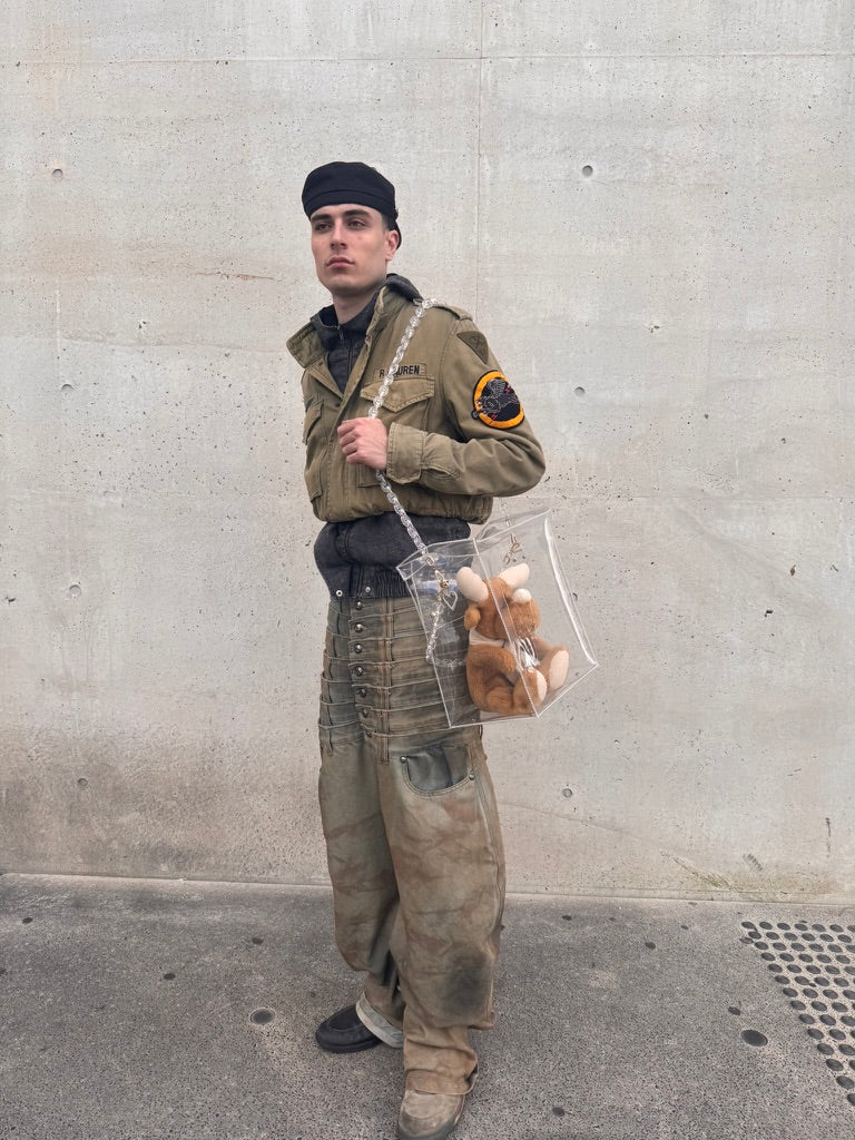 Person in military-style outfit holding a transparent bag with stuffed animals against a concrete wall.