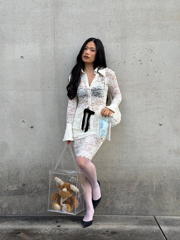 Woman in a white outfit holding a transparent bag with a stuffed animal against a concrete wall.
